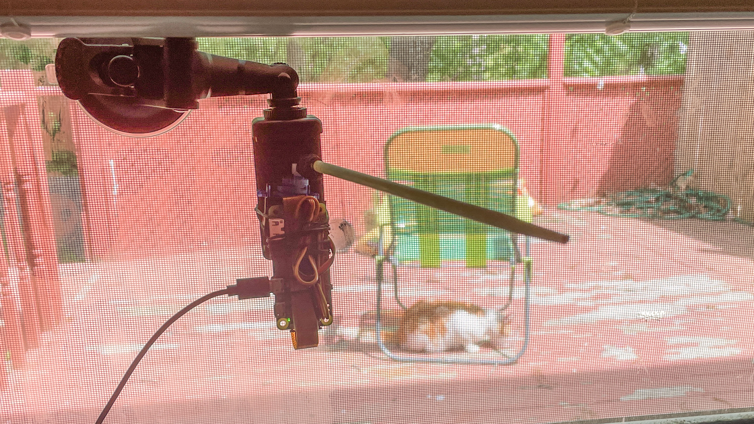 CatBot mounted on the window viewing a orange-and-white cat feeding on the porch.